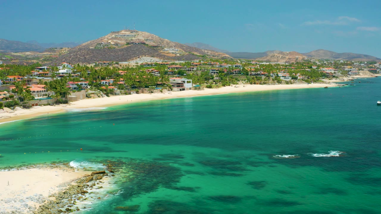 Coastal aerial with calm water, sandy beach, and hillside backdrop.