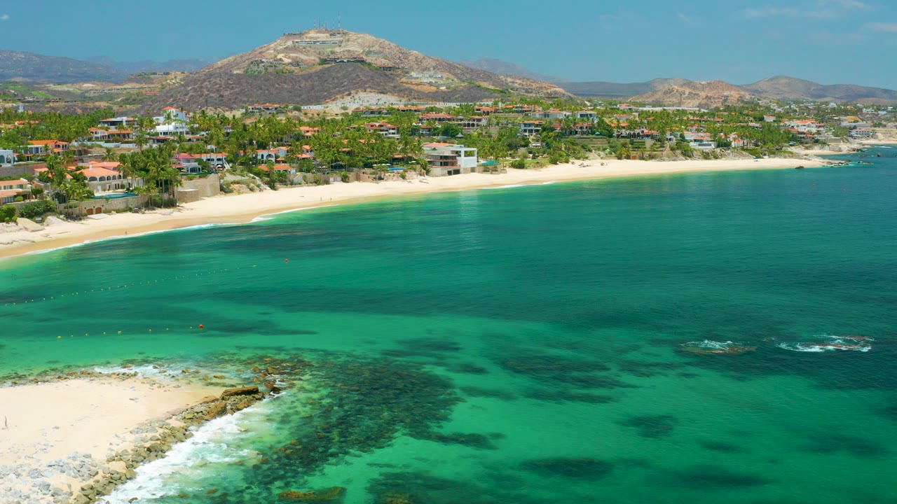 Aerial view over Palmilla Beach in Baja California Sur, with green ocean in front, pale sand and shoreline buildings beyond it, dry mountains in the background, and no visible people.