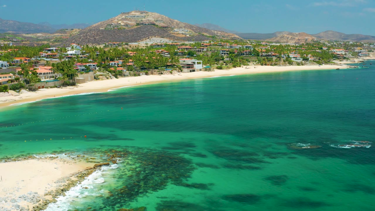 Palmilla Beach aerial view with green water and darker reef areas in the foreground, beachfront homes and palms along the sand, dry mountains behind, and no visible people.