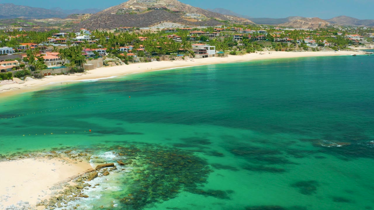 Aerial frame over Palmilla Beach, Baja California Sur, Mexico, with turquoise water, darker reef patches, pale sand, shoreline buildings, dry hills, and no visible people.