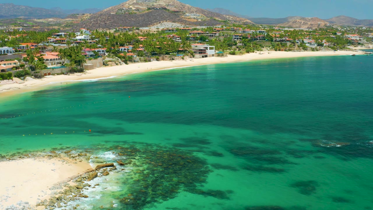 High aerial view at Palmilla Beach showing aqua water in the foreground, dark underwater patches, a narrow sand beach, palm-lined buildings, hills, and no visible people.