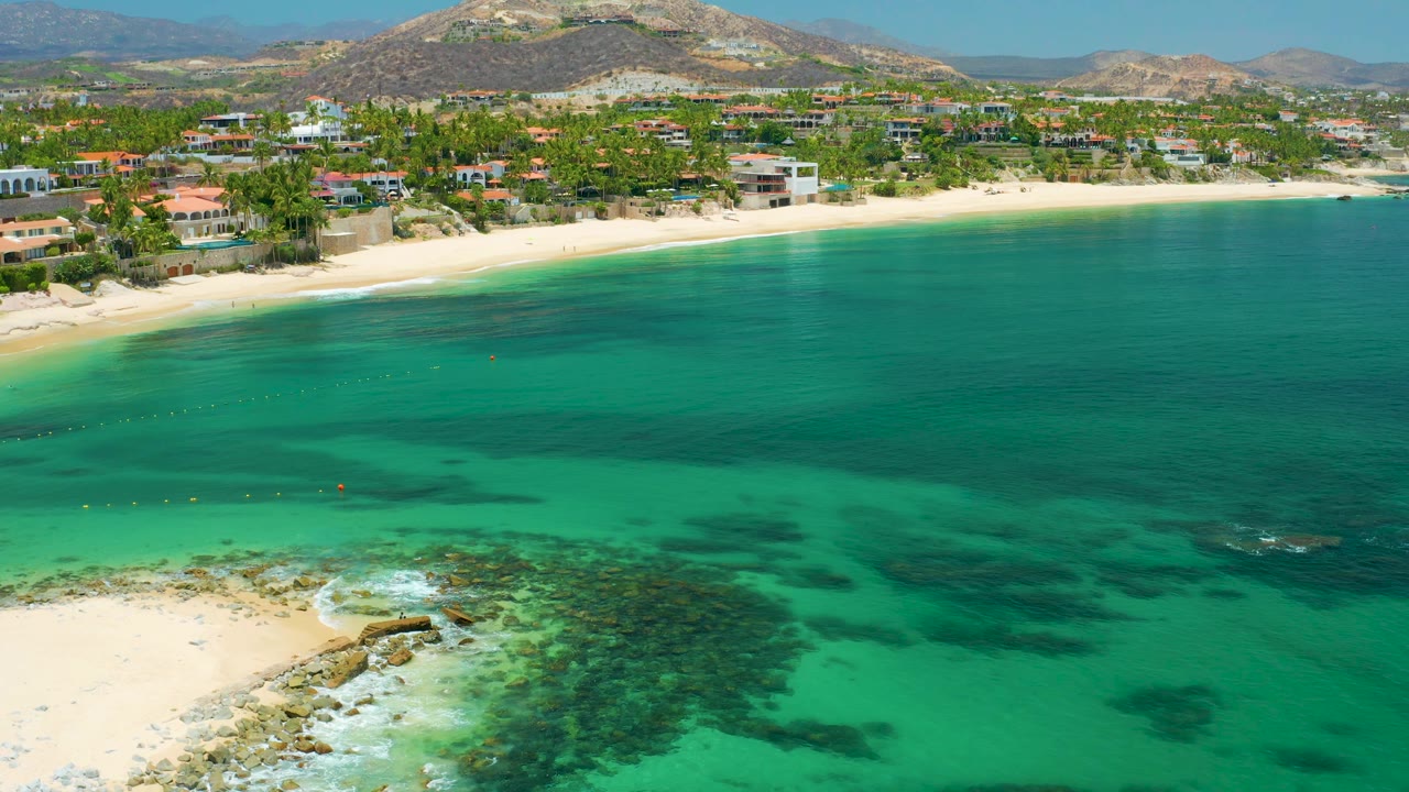 Wide drone frame from Palmilla Beach with green-blue water, a light sand shoreline, scattered palms and buildings, low brown hills behind them, and no visible people.