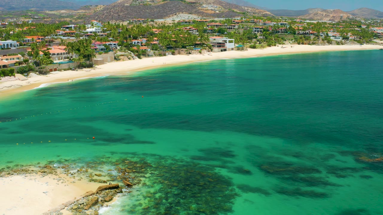 Second aerial frame over Palmilla Beach showing green-blue water with reef shadows, a pale sand strip, palm-lined beachfront homes, low arid hills, and no visible people.