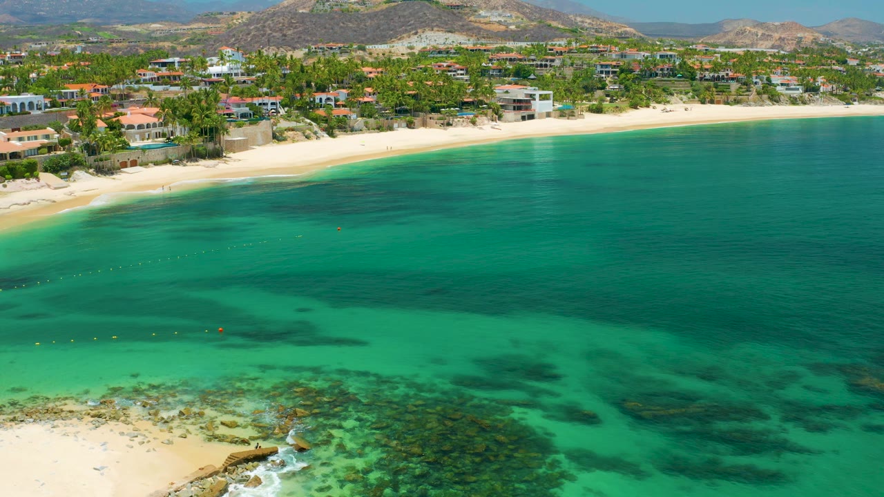 Aerial beach scene with shallow turquoise water, reef patches, small surf, homes, palms, and hills.