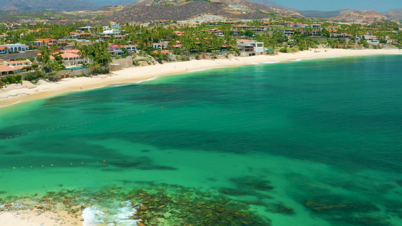 Opening aerial frame over Palmilla Beach, Baja California Sur, with rocky shallows at the lower edge, turquoise water, a pale beach, beachfront homes, palms, dry hills, and no visible people.