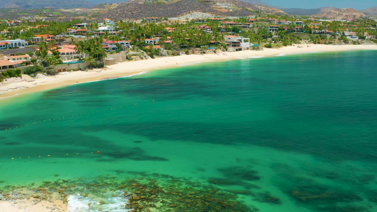 At 0:02, the aerial view shows Palmilla Beach from water to hillside, with darker underwater patches crossing the bay, a light sand strip, beachfront villas, palms, and no visible people.