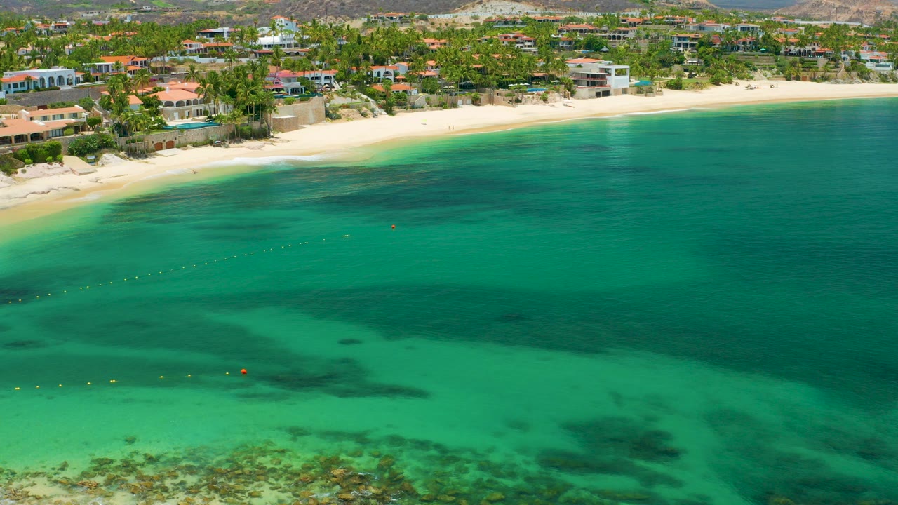 A drone frame over Palmilla Beach, Baja California Sur shows turquoise water in the foreground, a pale sand shoreline, beachfront homes, palms, and no visible people.