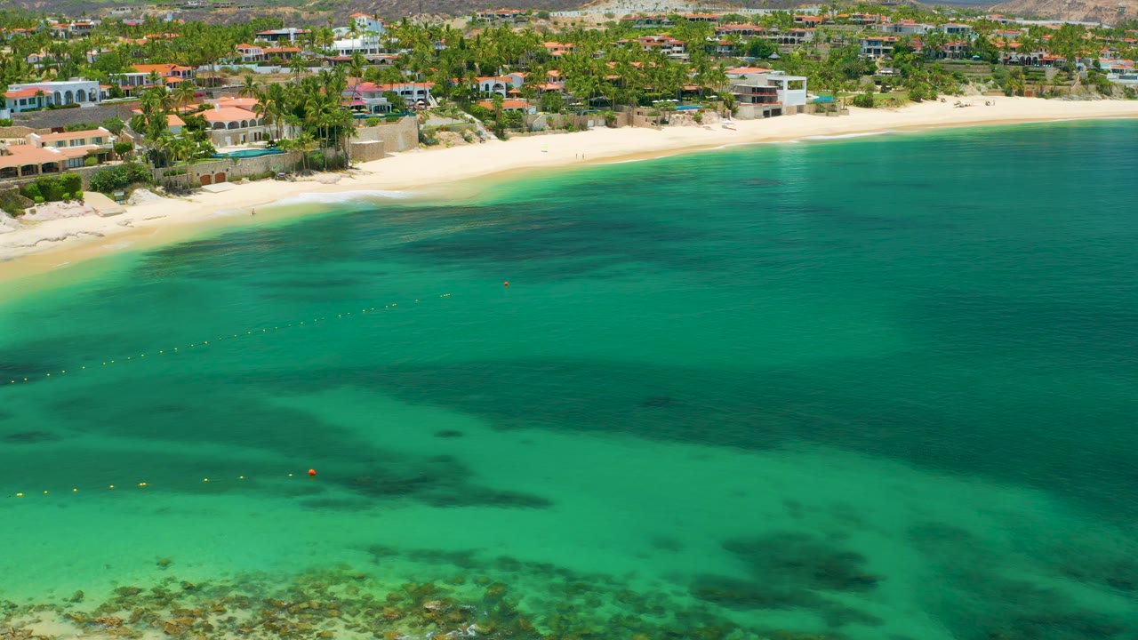 At 0:02, the aerial view holds on Palmilla Beach with calm turquoise water, a curved strip of sand, palm-lined homes, distant dry hills, and no visible people.