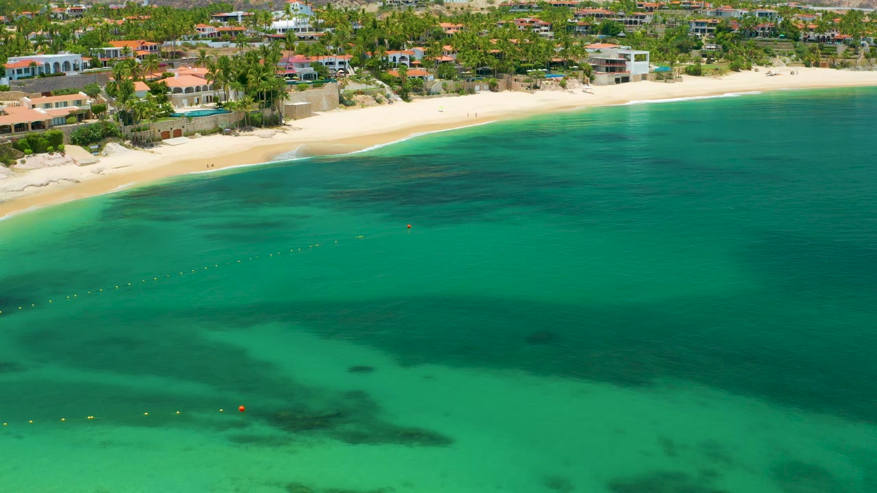 Drone-height view over green-blue water toward Palmilla Beach, showing darker reef or sea grass patches, a narrow sandy beach, waterfront homes, palms, and no visible people.
