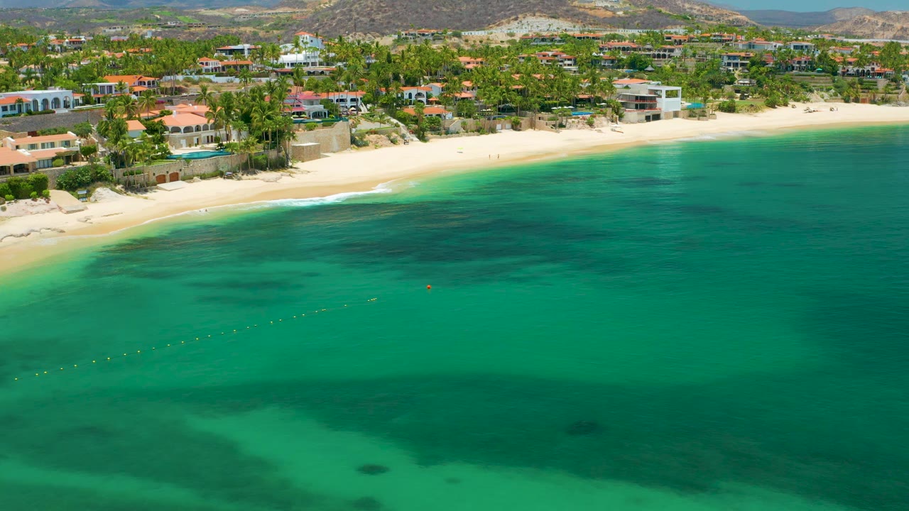 Aerial beach view with clear green water, pale sand, palms, buildings, and hills.