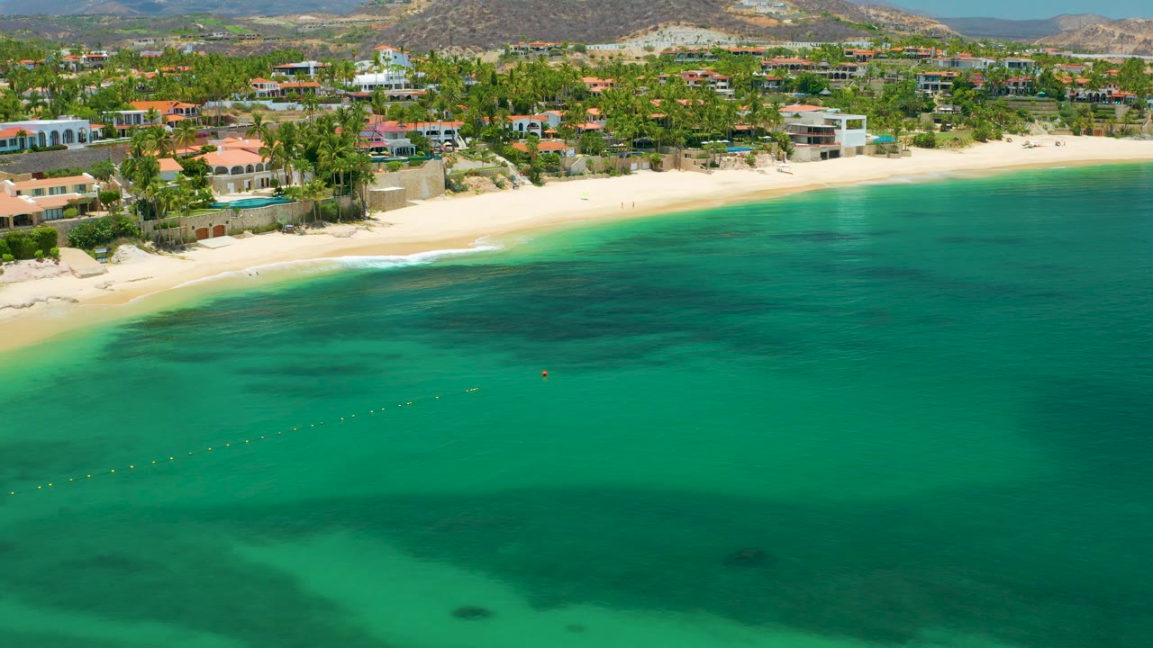 Wide Palmilla Beach aerial with a long curve of sand, calm turquoise water, palms and beachfront buildings behind the beach, low hills beyond, and no visible people.