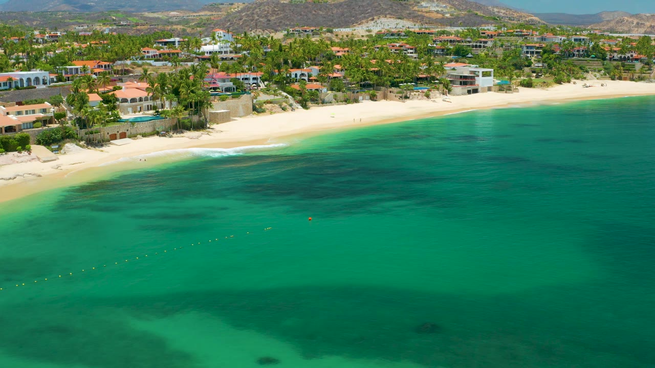 Aerial view of mild surf on a pale beach with green water and resort-side buildings.