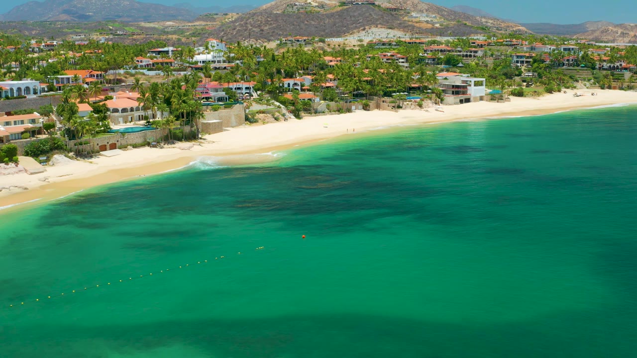 Aerial view of Palmilla Beach with a darker water patch near shore, bright sand, palm trees, large beachfront houses, low hills, and no visible people.