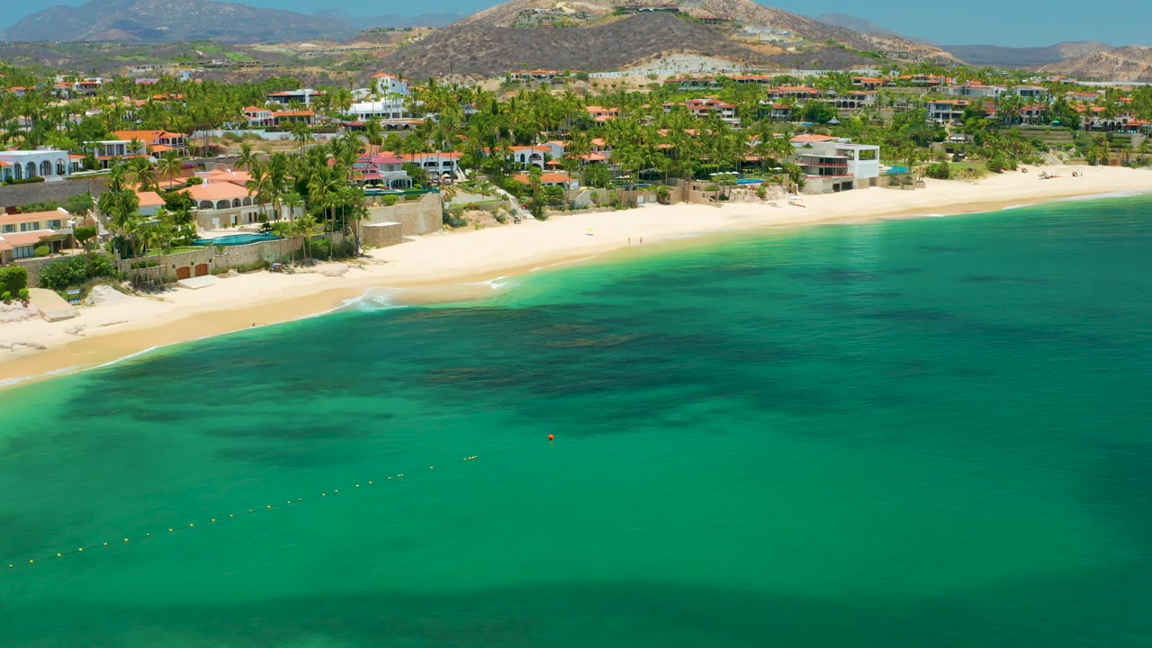 Palmilla Beach aerial frame showing a broad band of turquoise ocean, a lightly curved sandy shore, residential buildings among palms, dry hills behind, and no visible people.