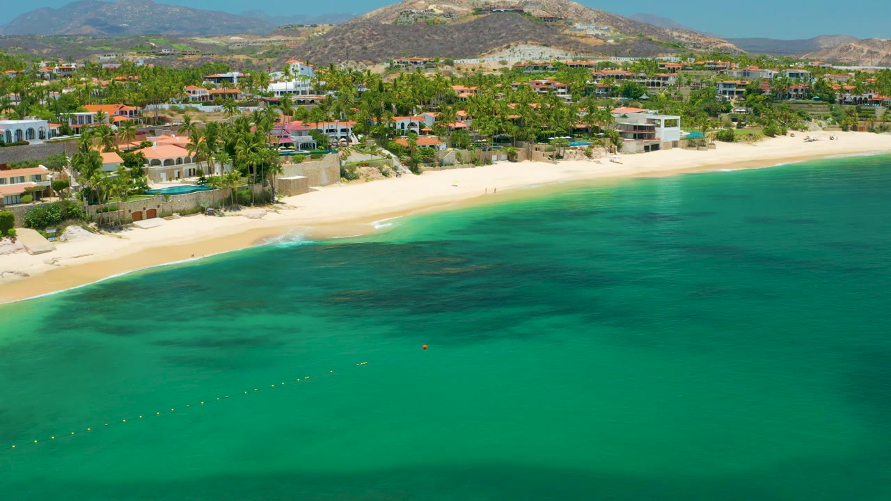 Aerial coastal view with beach, palms, houses, hills, and open water.