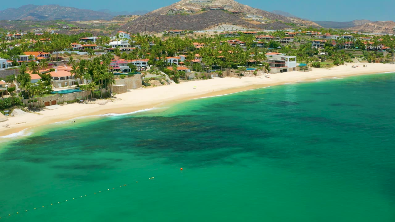 Wide aerial view of Palmilla Beach with turquoise shallows, a curving sand beach, clustered beachfront buildings, palms, distant hills, and no visible people.