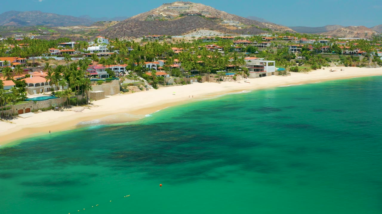 Aerial frame over Palmilla Beach in Baja California Sur with turquoise water below, a pale curved beach, resort buildings and palms behind it, dry hills in the distance, and no visible people.