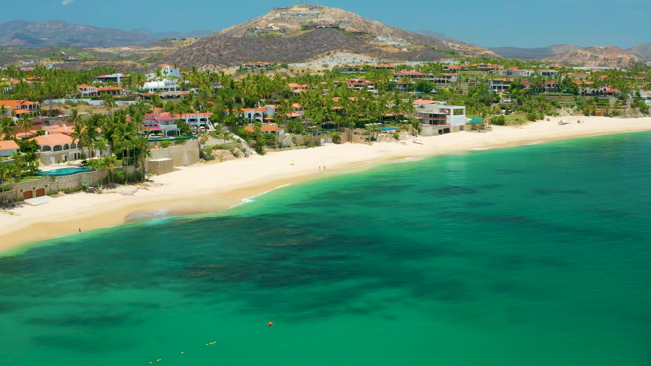 Palmilla Beach aerial frame showing a broad turquoise foreground, a narrow white-sand shoreline, low resort buildings and palms across the back of the beach, distant hills, and no visible people.