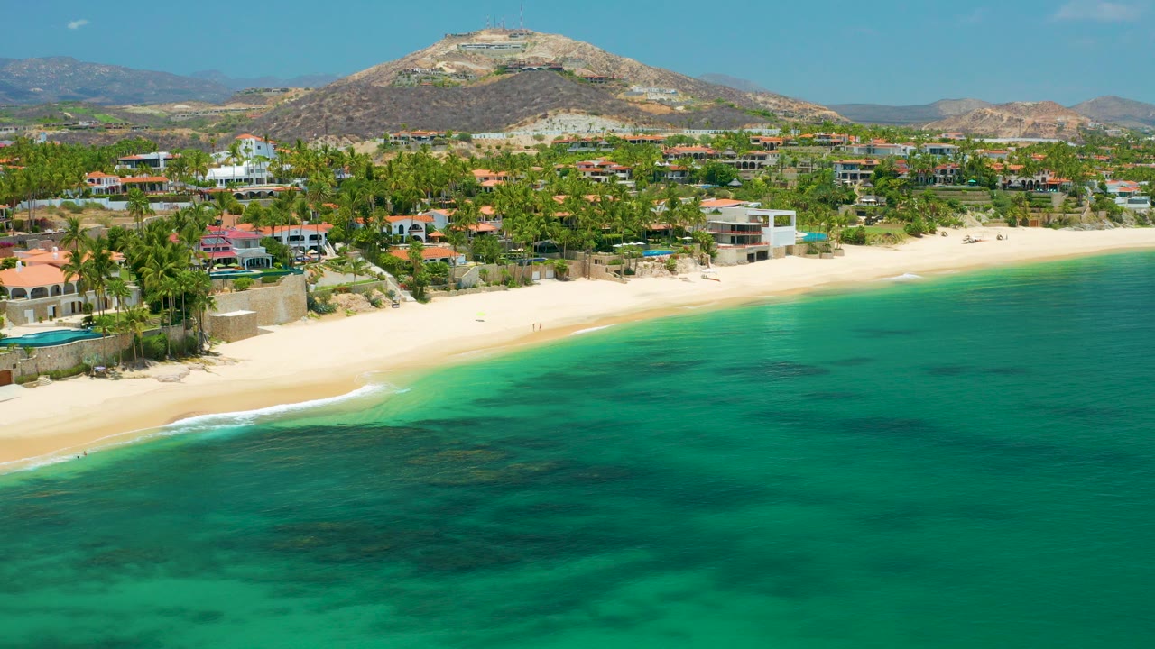 Palmilla Beach, Baja California Sur seen from above with turquoise ocean in the foreground, a curved sandy shore, palm-lined homes, dry hills, and no clearly visible people.