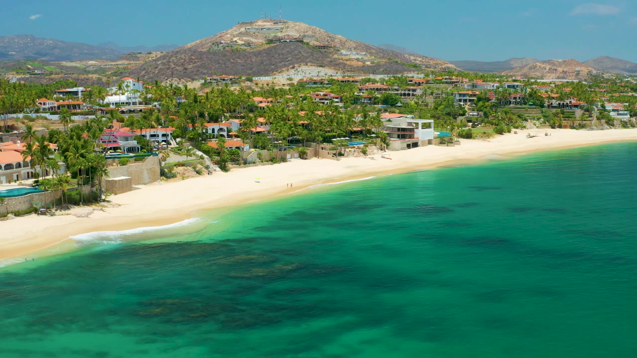 Drone view of Palmilla Beach with waves, sand, homes, palms, and mountains.