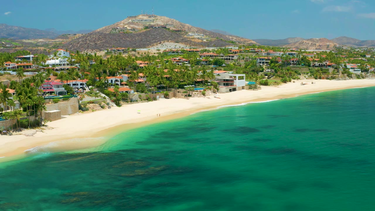 Wide coastal aerial with green water, sand beach, homes, palms, and mountain backdrop