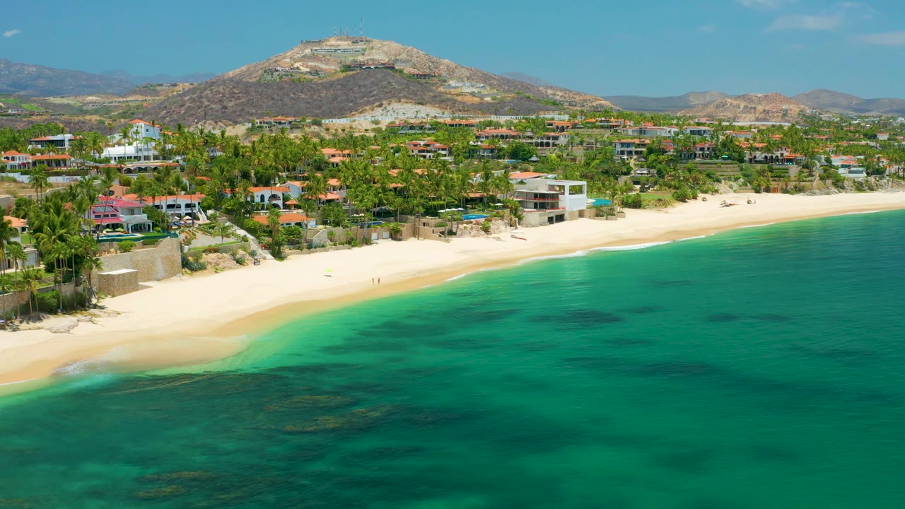 Aerial view at Palmilla Beach with the clear water pushed farther across the foreground, a thin surf line on the sand, homes and palms along the coast, hills beyond, and no visible people.