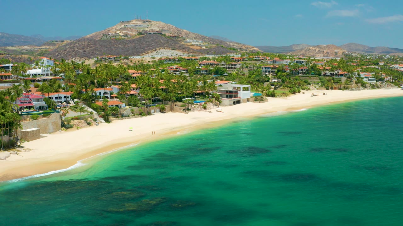 Opening aerial frame over Palmilla Beach in Baja California Sur, with turquoise sea in front, a pale sand shoreline, beachfront buildings, palms, dry hills, and no visible people.