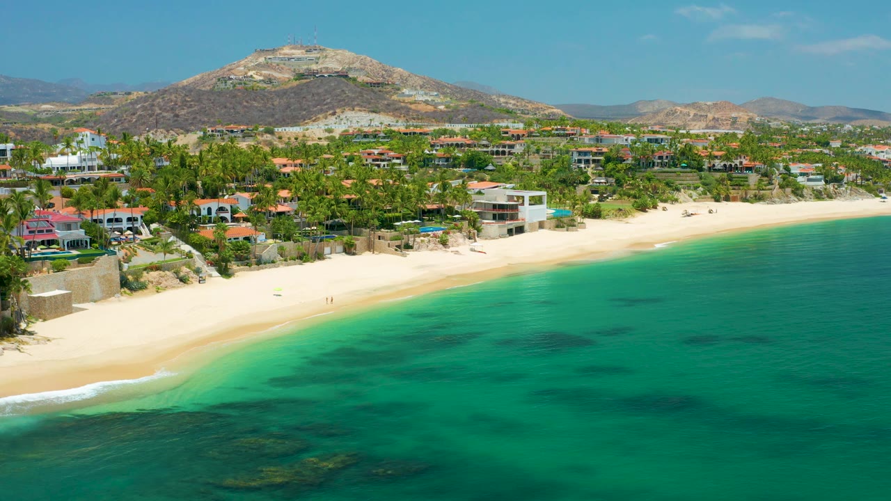 Aerial view along a pale beach with small surf, green water, palms, buildings, and hills.