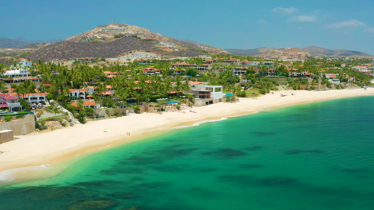 Drone view of a sandy beach beside green-blue water and palm-lined residences.