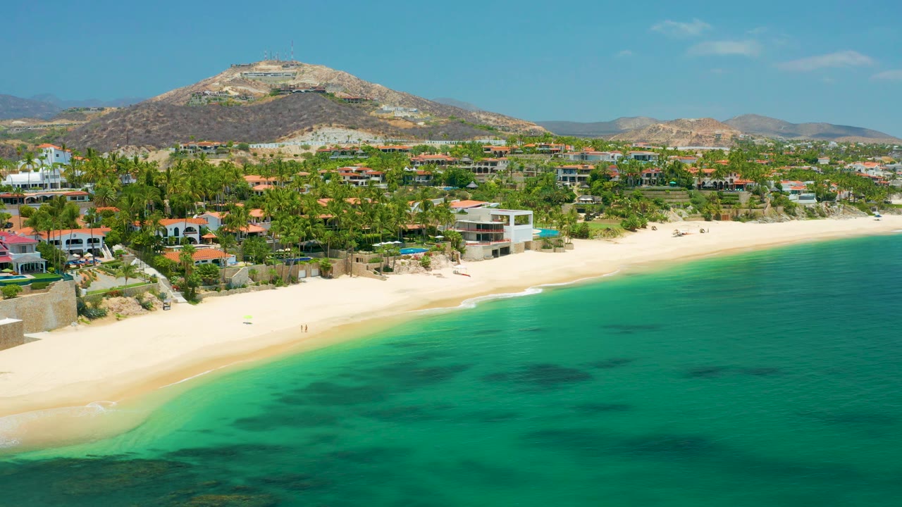 Wide aerial frame of Palmilla Beach with turquoise sea filling the right foreground, pale sand along the left, coastal homes and palms behind it, hills beyond, and no visible people.