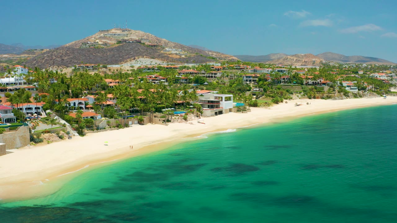 Aerial beach scene with pale surf edge, coastal houses, palms, and rocky hills.
