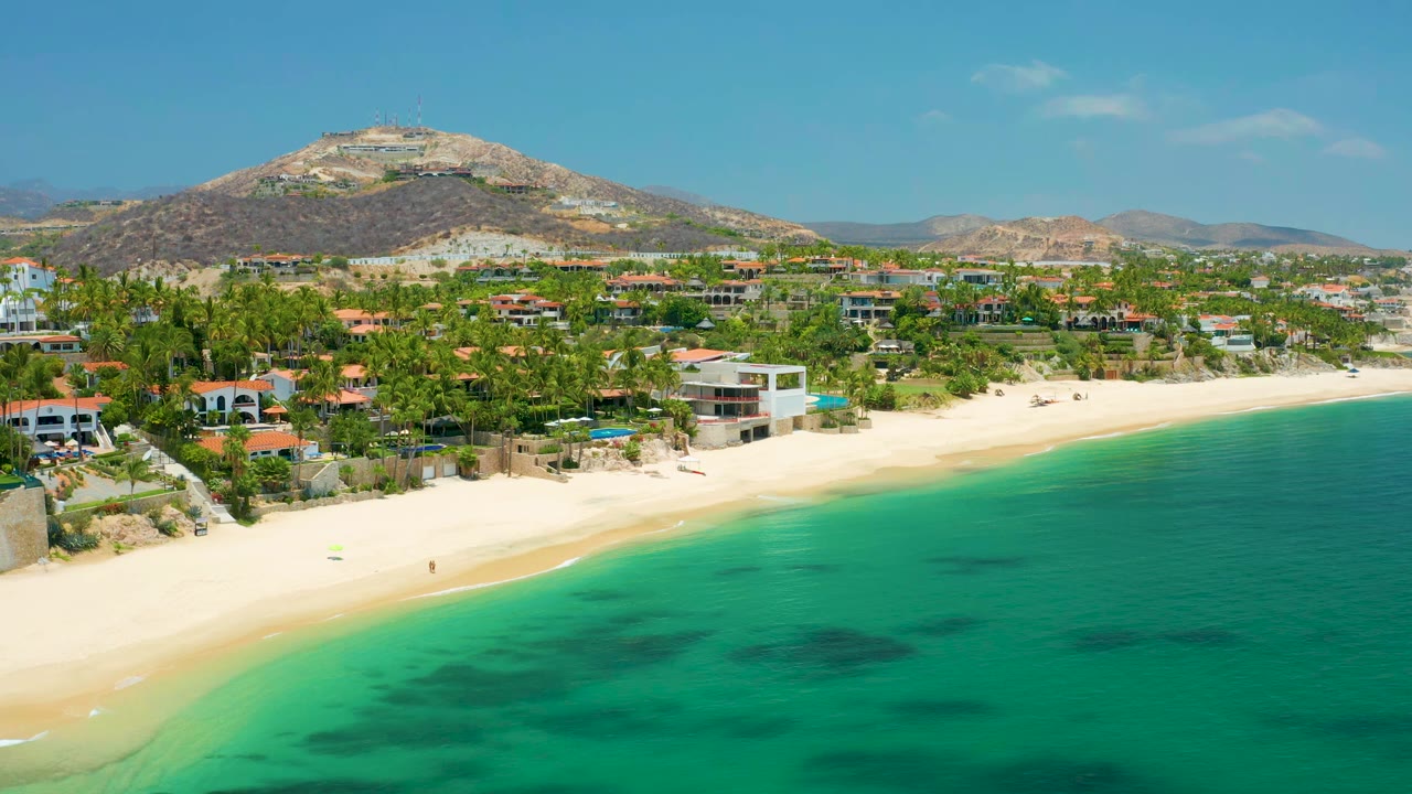 Palmilla Beach aerial view one second into the clip, showing more of the shoreline arc, green-blue shallows, beachfront houses, palm trees, distant hills, and no visible people.