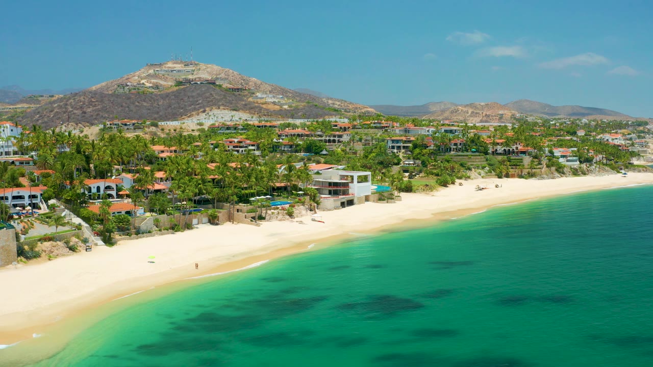 Aerial view at Palmilla Beach with the sea occupying the lower right, a bright sand strip along the coast, homes among palms, a rounded mountain behind town, and no visible people.