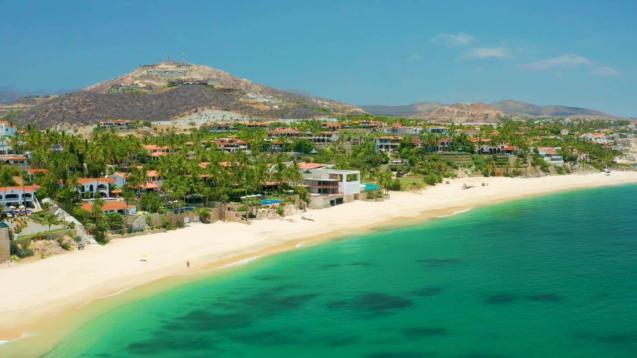 Wide aerial coastline with turquoise water, empty-looking sand, palms, houses, and dry hills.