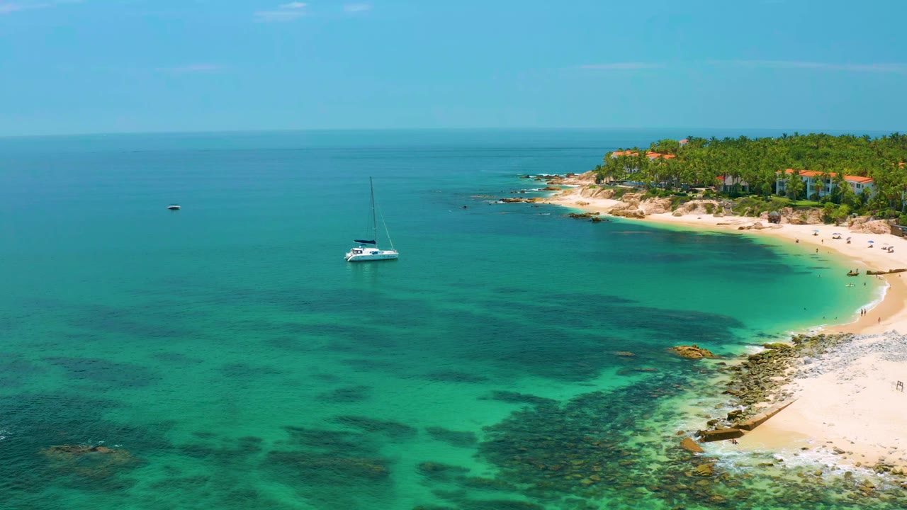Palmilla Beach aerial view with the sailboat left of the beach curve, shallow reef patches visible through the water, green trees on the point, and no visible people.