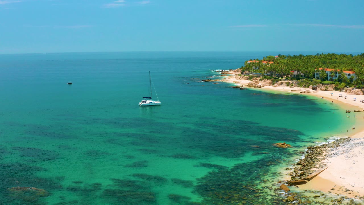 Aerial view of Palmilla Beach with a white sailboat in blue-green water, a pale curved shoreline to the right, dark underwater patches, distant palms and low buildings, and no visible people.