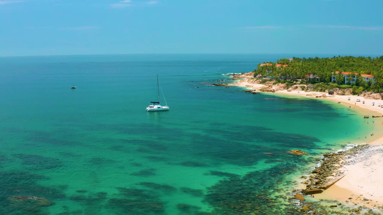 Palmilla Beach remains at frame right as the white sailboat sits in the midground over turquoise water, with reef-like dark patches below, low buildings beyond the palms, and no visible people.