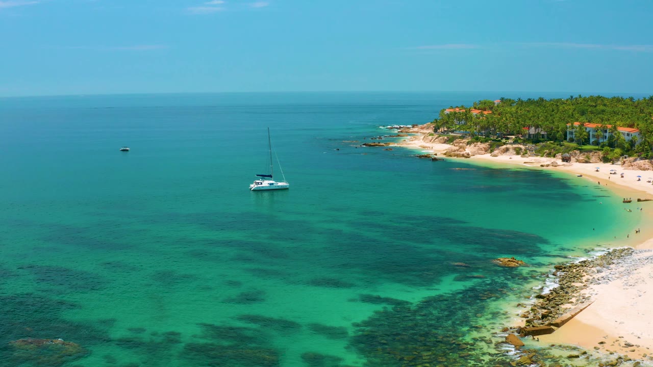 Aerial frame from Palmilla Beach shows the sailboat slightly left of center, open blue water across the left side, the sandy beach and palms to the right, and no visible people.