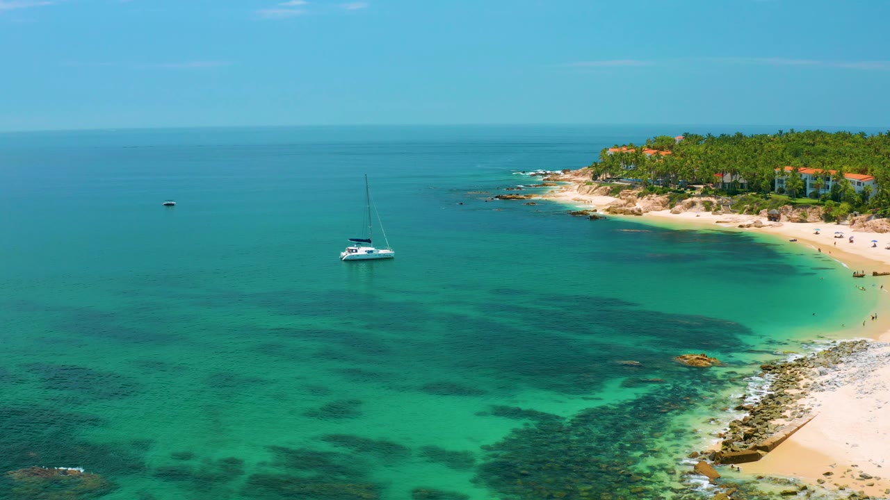 Aerial coastline view with sailboat, clear water, beach, and palms.
