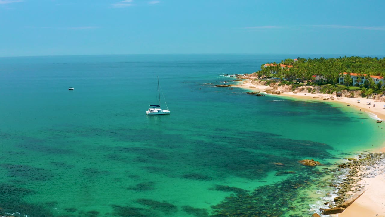 Aerial view over Palmilla Beach in Baja California Sur with turquoise water, a white sailboat offshore, sandy shoreline at right, palms in the distance, and no visible people.