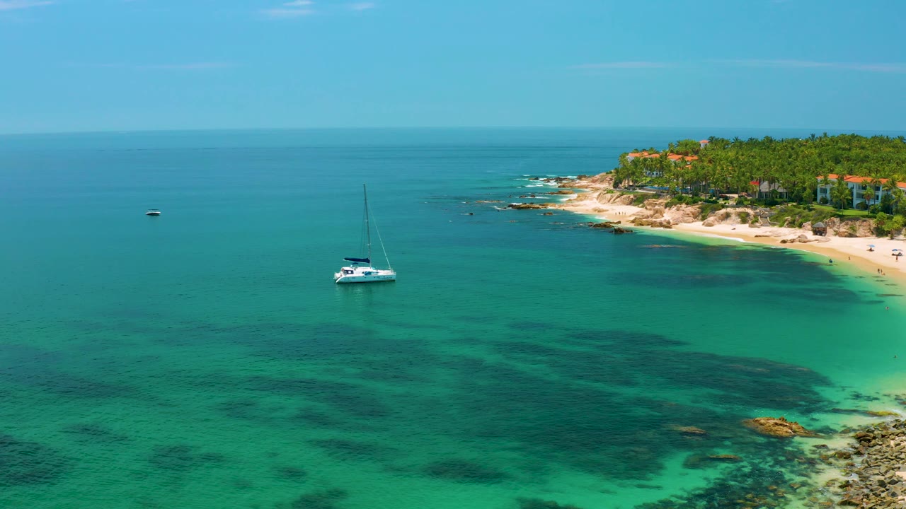 Wide aerial view of a white yacht off Palmilla Beach, Baja California Sur, with dark reef areas under the water, a rocky sandy shoreline on the right, and small buildings above the beach.