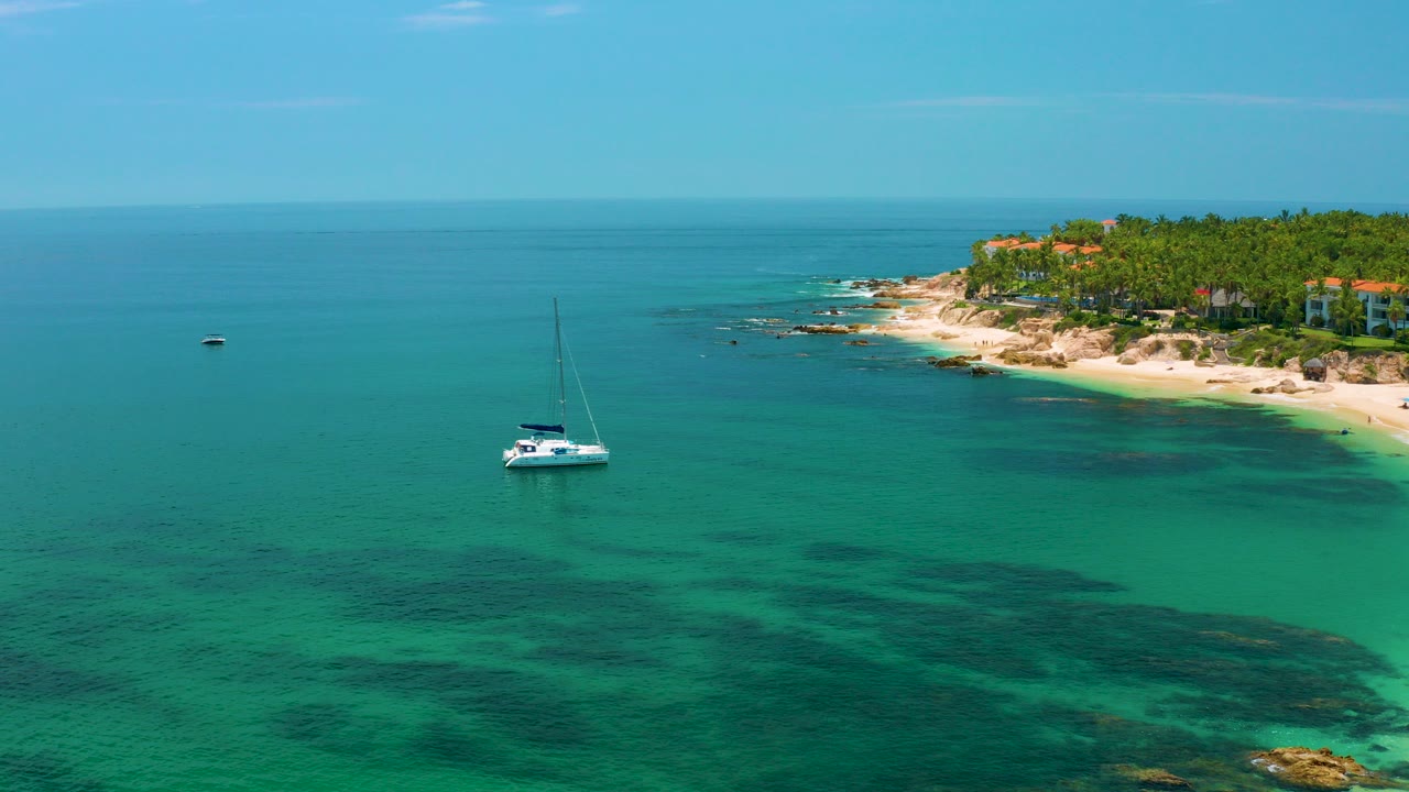 A white sailboat floats offshore at Palmilla Beach, Baja California Sur, with dark underwater patches, a sandy palm-lined coast to the right, and no visible people.