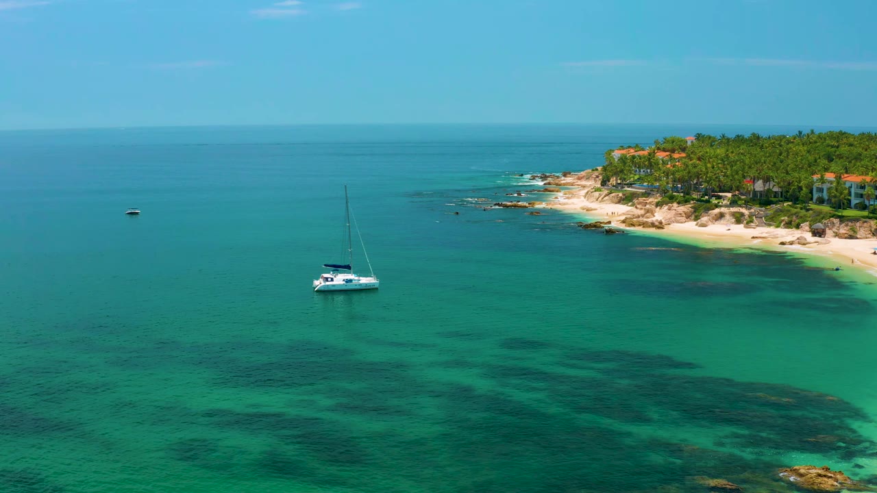 The sailboat sits left of the Palmilla Beach shoreline as turquoise water fills the foreground, palm trees and pale sand line the right side, and no visible people appear.