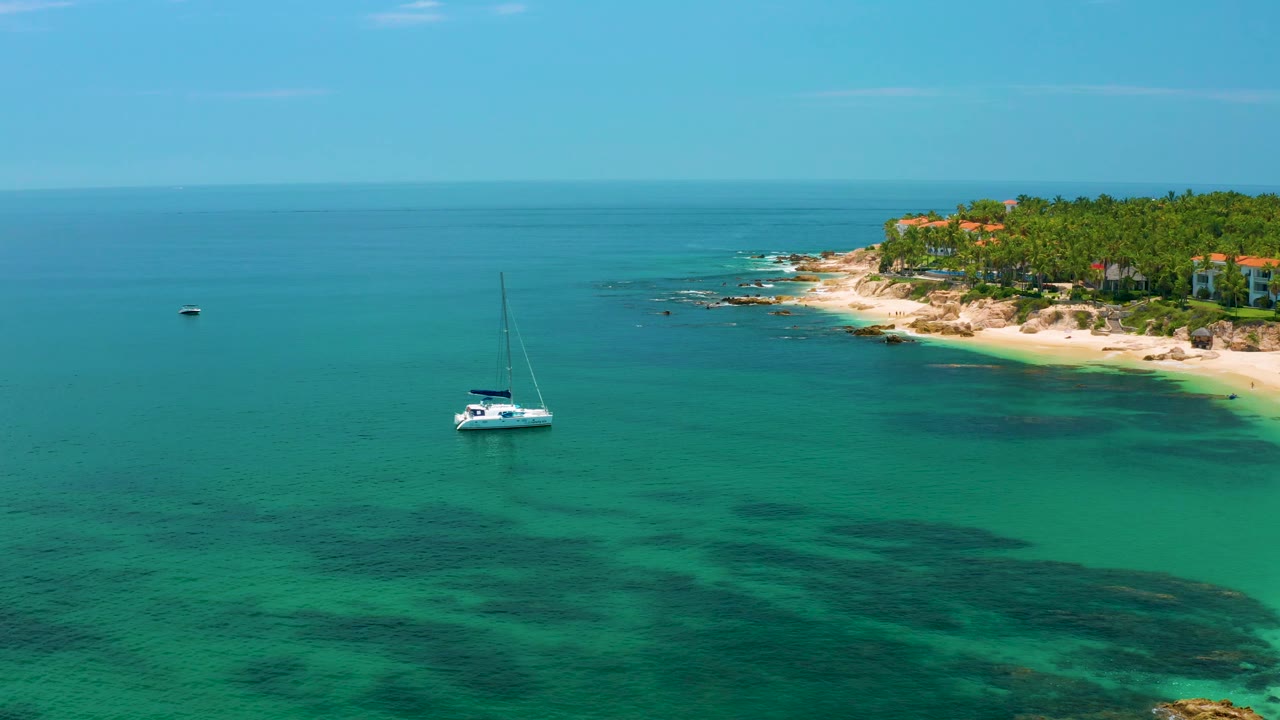 Aerial view over Palmilla Beach shows the sailboat below the horizon, the rocky sandy coast at right, green palms behind it, and no visible people in the frame.