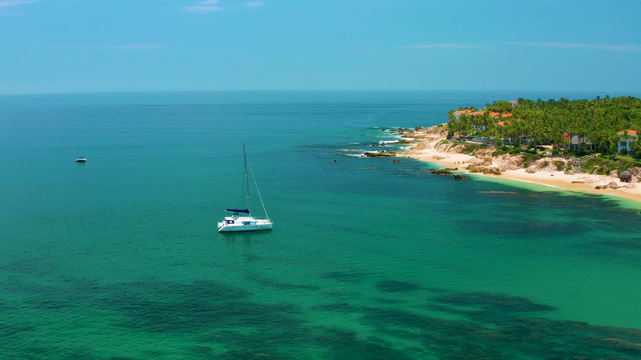 White sailboat floats on green water off Palmilla Beach, Baja California Sur, with dark underwater patches and a palm-lined rocky shore in the distance, no visible people.