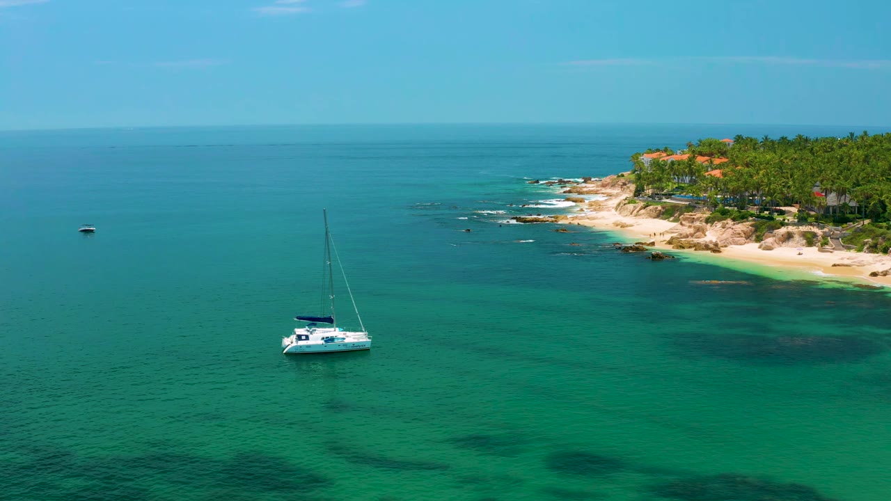 Anchored white catamaran remains in clear green-blue water below the Palmilla Beach coastline, with palm trees and rocky sand in the upper right and no visible people.