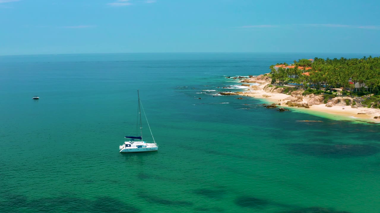 White catamaran sits slightly right of center in shallow teal water, with darker reef-like patches beneath the surface, Palmilla Beach shoreline behind it, and no visible people.