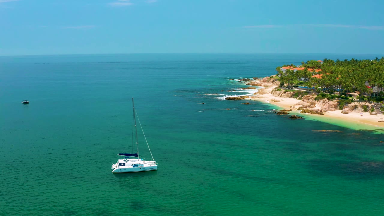 Aerial frame over Palmilla Beach showing the catamaran slightly left of center, a broad blue horizon, shallow green water, pale beach, trees, and no visible people.