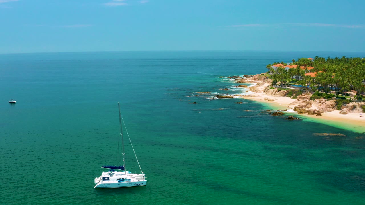 A white sailboat sits low in the frame on turquoise water near the rocky Palmilla Beach shoreline in Baja California Sur, with palms and red roofs in the distance and no visible people.