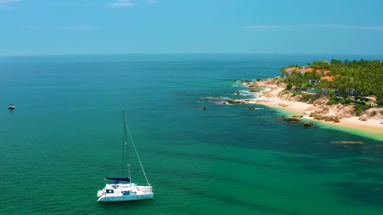 A white sailboat appears over darker green water as the rocky Palmilla Beach edge and palm-covered land remain in the upper right, with no visible people in the frame.